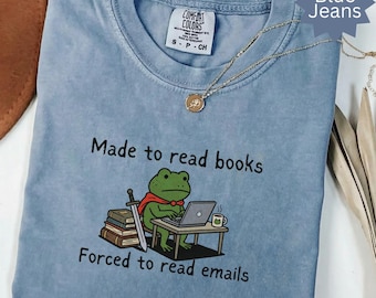Może przedstawiać: Jasnoniebieska koszulka Comfort Colors z napisem "Made to read books, Forced to read emails". Koszulka przedstawia kreskówkową żabę w pelerynie i używającą laptopa. Widoczny jest również złoty naszyjnik z wisiorkiem.