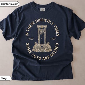 May include: A navy blue short-sleeved t-shirt featuring a cream-coloured graphic of a guillotine flanked by roses. The text reads: IN THESE DIFFICULT TIMES EST. 1792 SOME CUTS ARE NEEDED. The shirt has a crew neckline and a Comfort Colors brand label.