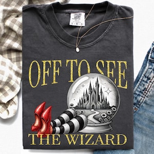 Könnte beinhalten: Ein anthrazitfarbenes T-Shirt mit dem goldglitzernden Schriftzug "OFF TO SEE THE WIZARD". Die Grafik zeigt eine schwarz-weiße Illustration eines Schlosses in einer Kristallkugel, begleitet von roten Schuhen und schwarz-weiß gestreiften Beinen.