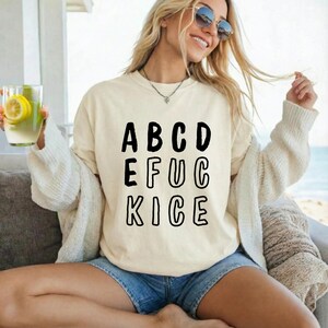 May include: Cream-colored sweatshirt with the word "ABCDEFUCKICE" in black block letters. The person in the image is wearing sunglasses and denim shorts. She is holding a glass of lemonade.