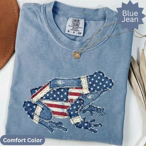 May include: Blue jean Comfort Colors t-shirt featuring an appliqued frog design with the American flag. The shirt has a crew neck and short sleeves. A gold necklace with a pendant is draped on the shirt.