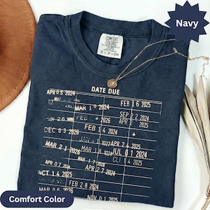 May include: Navy blue Comfort Colors t-shirt with a gold necklace. The shirt features a printed chart with the words "DATE DUE" and various dates. The shirt is a size small and has a crew neck.