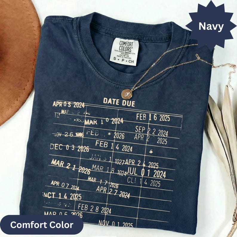 May include: Navy blue Comfort Colors t-shirt with a gold necklace. The shirt features a printed chart with the words "DATE DUE" and various dates. The shirt is a size small and has a crew neck.