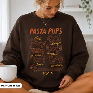 Peut inclure: Un sweat-shirt à col rond marron chocolat foncé avec une impression graphique portant le texte PASTA PUPS et des illustrations de bouledogues français stylisés en diverses formes de pâtes comme fusilli, lasagna, fettuccine, cavatappi, penne, macaroni et spaghetti.