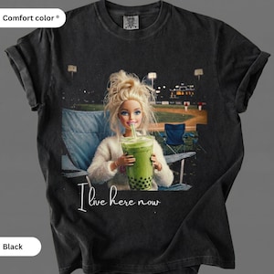 May include: Black Comfort Colors t-shirt featuring a graphic of a doll drinking a green beverage with the text "I live here now." The doll is seated in a chair with a baseball field in the background.
