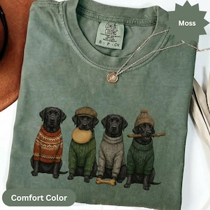 May include: A moss green Comfort Colors t-shirt featuring a graphic of four black Labrador Retrievers wearing sweaters and hats. One dog holds a bone, another a frisbee, and another a stick. A silver necklace with a pendant is also visible.