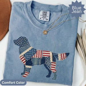 May include: A light blue Comfort Colors t-shirt featuring a patchwork dog design with American flag patterns. The shirt has a crew neck and short sleeves. A gold necklace with a pendant is draped across the shirt. The words "Blue Jean" and "Comfort Color" are visible.