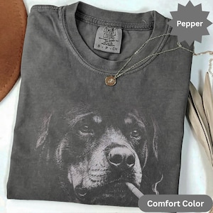 Pode incluir: Camiseta Comfort Colors cor pimenta com um gráfico em preto e branco de um cachorro fumando um cigarro. A camiseta tem gola redonda e mangas curtas. Um colar de prata com um pingente está sobre a camiseta.