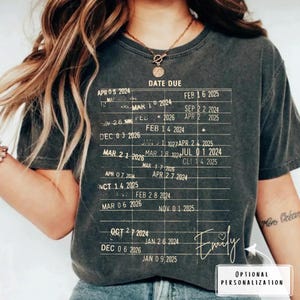 May include: Dark gray t-shirt with a printed chart design featuring dates and the text "DATE DUE." The shirt includes the name "Emily" in cursive and the words "OPTIONAL PERSONALIZATION" in a white box.
