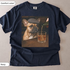 Könnte beinhalten: Ein marineblaues T-Shirt mit einem Grafikdruck eines falbfarbenen Französischen Bulldoggen, der eine graue Schiebermütze trägt. Der Hund befindet sich neben einem Glas, das aus einer Flasche mit einer bernsteinfarbenen Flüssigkeit gefüllt wird.