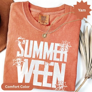 Op de afbeelding: Een yam-kleurig Comfort Colors T-shirt met de tekst "SUMMER WEEN" in het wit, samen met spinnenwebafbeeldingen. Een gouden ketting met een hanger is over het shirt gedrapeerd.
