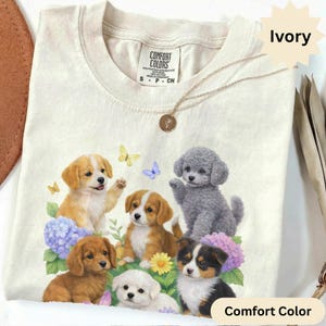 Peut inclure: T-shirt ivoire Comfort Colors avec un motif coloré de six chiots entourés de fleurs et de papillons. Les chiots sont de différentes races et couleurs, dont dorés, bruns, blancs et gris. Le t-shirt a un col rond et des manches courtes.