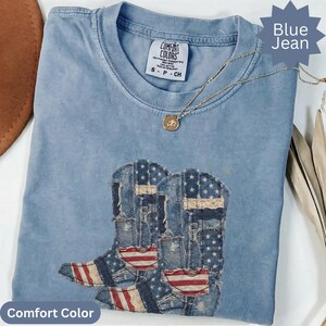 May include: A blue jean Comfort Colors t-shirt with a graphic of two cowboy boots decorated with the American flag. The shirt has a crew neck and short sleeves. A gold necklace with a pendant is draped across the shirt.