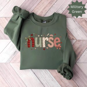 May include: A military green sweatshirt featuring the word "nurse" in a patchwork design. The letters are red, pink, and green with heart and bow accents. The sleeve cuffs are folded up.