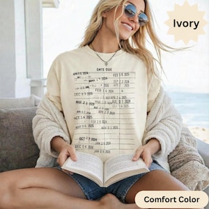 May include: Ivory-colored Comfort Color t-shirt with a vintage library card design printed on the front. The graphic features a grid with the words "DATE DUE" and various dates. The person is wearing sunglasses and holding a book.