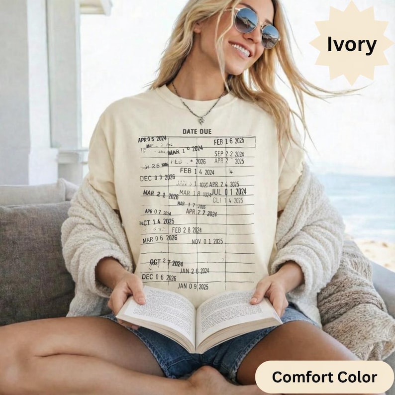 May include: Ivory-colored Comfort Color t-shirt with a vintage library card design printed on the front. The graphic features a grid with the words "DATE DUE" and various dates. The person is wearing sunglasses and holding a book.