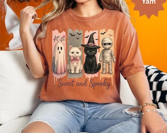 Pode incluir: Uma camiseta Comfort Color cor inhame com um gráfico temático de Halloween. O gráfico inclui um fantasma, um gato com um laço rosa, um gato preto com um chapéu de bruxa e uma múmia. O texto "Sweet and Spooky" está abaixo.