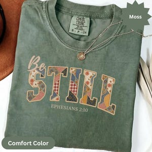 Puede incluir: Una camiseta de manga corta color verde musgo con cuello redondo. El frente presenta el texto "Be STILL" en un patrón de patchwork naranja, azul y blanco, con "EPHESIANS 2:10" impreso debajo. Una fina cadena dorada descansa sobre la tela.