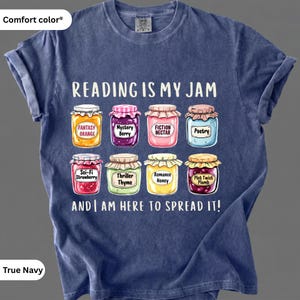 Puede incluir: Camiseta True Navy Comfort Colors con el texto "READING IS MY JAM" e ilustraciones de tarros de mermelada etiquetados con géneros de libros como "Fantasy Orange" y "Mystery Berry". La camiseta también dice "AND I AM HERE TO SPREAD IT!"