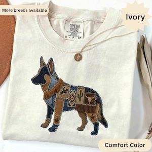 Op de afbeelding: Een ivoorkleurig t-shirt met een Comfort Colors-label en een afbeelding van een Duitse herder. Het hondenontwerp is samengesteld uit verschillende stoffen patches, waaronder denim, bruin suède en franjes, met iconen van een cowboy, cactus en schedel.