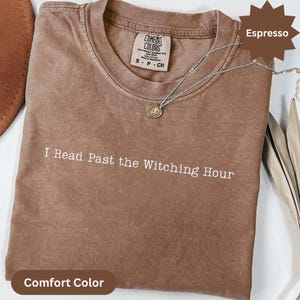 Puede incluir: Camiseta Comfort Colors color Espresso con la frase "I Read Past the Witching Hour" impresa en blanco. Un collar plateado con un colgante descansa sobre la camiseta doblada.
