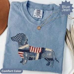 May include: Blue jean Comfort Colors t-shirt with a dachshund dog design. The dog is made of denim patches with an American flag theme. A gold necklace with a pendant is draped across the shirt.