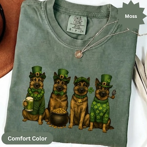 May include: A moss green Comfort Colors t-shirt featuring four German Shepherds dressed for St. Patrick's Day. Each dog wears a green hat, and some have accessories like a beer mug, pot of gold, and shamrocks. A silver necklace is draped across the shirt.