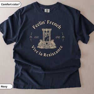 May include: A navy blue short-sleeved t-shirt featuring a cream-coloured graphic of a guillotine flanked by roses. The text reads Feelin' French, EST. 1792, and Vive la Resistance, accented with small crescent moon and star symbols around the central design.