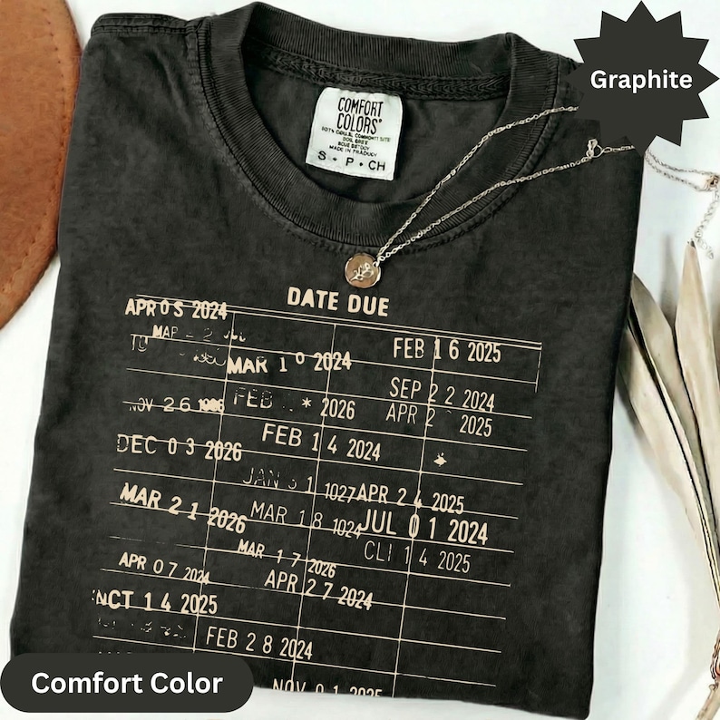 May include: Graphite-colored Comfort Colors t-shirt with a printed "Date Due" design. The design includes dates and a small Comfort Colors tag. A silver necklace with a pendant is draped over the shirt.