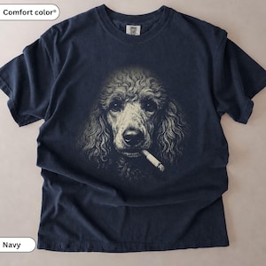 May include: A navy blue short-sleeve t-shirt featuring a graphic print of a curly-haired poodle with a cigarette in its mouth. The shirt has a crew neckline and a Comfort Colors brand label on the inside collar. The poodle illustration is in shades of cream and off-white, contrasting against the dark fabric of the garment.