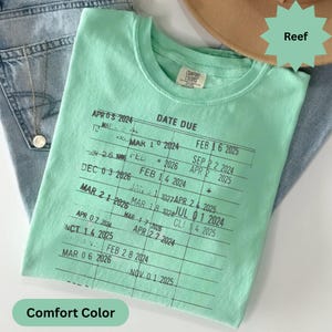 May include: A mint green Comfort Colors t-shirt with a printed "Date Due" design. The design features a grid with dates and the word "Reef" on a green starburst. The shirt is folded on top of blue jeans.