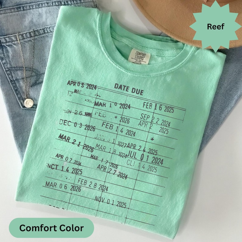 May include: A mint green Comfort Colors t-shirt with a printed "Date Due" design. The design features a grid with dates and the word "Reef" on a green starburst. The shirt is folded on top of blue jeans.