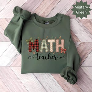 May include: A military green sweatshirt with the words "MATH teacher" in a decorative font. The word "MATH" is designed with plaid, polka dot, and patterned letters. The sweatshirt has long sleeves and a crew neck.