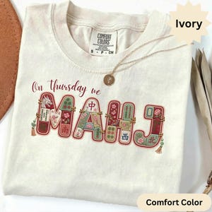 May include: An ivory t-shirt featuring the text On Thursday we MAHJ in a maroon font. The letters are filled with colorful illustrations of mahjong tiles, including floral patterns, a dragon, and Chinese characters, accented with gold tassels.