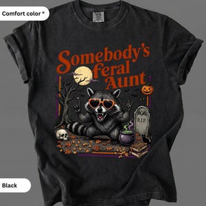 Pode incluir: Uma t-shirt preta de manga curta com um gráfico de um guaxinim a usar óculos de sol cor de laranja em forma de coração. O texto diz Somebody's feral Aunt. O design inclui uma lua cheia, uma árvore nua, um crânio, uma lápide e um caldeirão.
