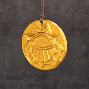 Peut inclure: Un pendentif doré avec un motif détaillé, suspendu par un cordon marron. Le pendentif présente une forme centrale en forme d'œil entourée d'un motif en étoile. Le pendentif est rond et a une surface texturée.