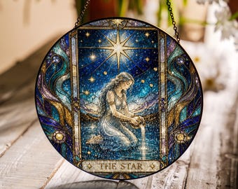 May include: A circular stained glass suncatcher featuring The Star tarot card design. It depicts a kneeling figure pouring water into a pool under a large golden star and smaller stars. The border has blue, teal, and gold swirling patterns.