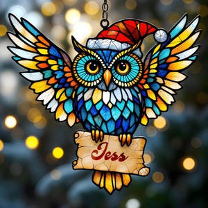 Puede incluir: Adorno de búho de vidriera con alas extendidas, con colores vibrantes de azul, amarillo, naranja y rojo. El búho lleva un gorro de Papá Noel y sostiene un pergamino con el nombre "Jess". Un artículo decorativo navideño.