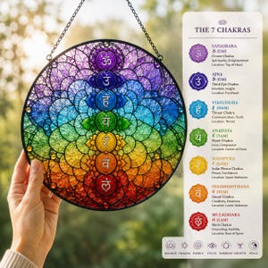 May include: A circular stained glass suncatcher featuring the seven chakra symbols in a vertical rainbow gradient from purple to red. The piece is framed in black metal and includes a hanging chain. A white informational card lists the 7 chakras.