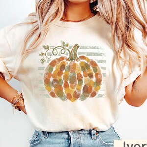 May include: An ivory short-sleeve t-shirt featuring a central graphic of a pumpkin composed of various colored fingerprints in shades of orange, green, and brown. The pumpkin design is set against a background of horizontal light green stripes.