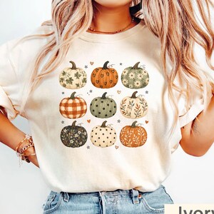May include: An ivory t-shirt featuring a graphic print of nine pumpkins arranged in a grid. The pumpkins display various patterns including plaid, polka dots, floral motifs, and heart shapes, interspersed with small star and heart icons.
