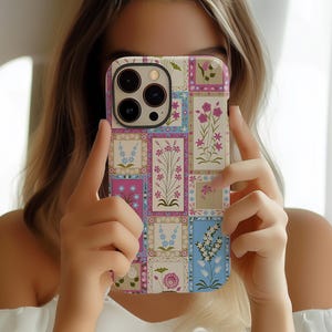 Puede incluir: Una funda para teléfono con un patrón floral en tonos de rosa, azul y verde. La funda tiene un diseño de patchwork con cuadrados y rectángulos de diferentes tamaños. La lente de la cámara es visible en la parte posterior de la funda.