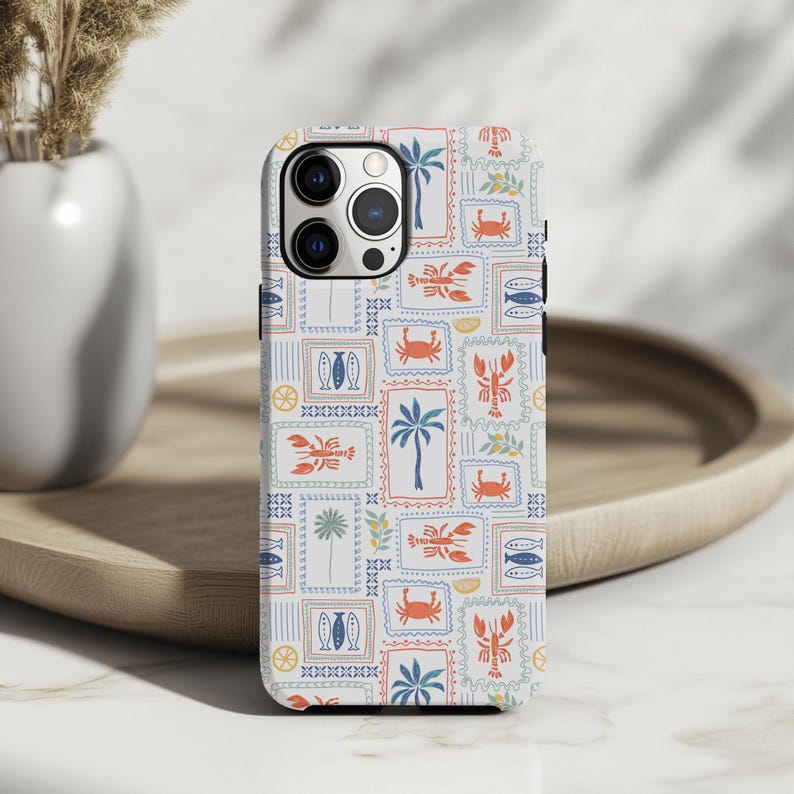 May include: A phone case with a repeating pattern of postage stamp-like designs. Each stamp features illustrations of palm trees, fish, crabs, and lobsters in shades of blue, orange, and green on a white background. The case is on a wooden tray.