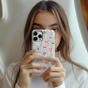 May include: A white phone case with a colorful, postage stamp-style design featuring crabs, fish, palm trees, and other nautical elements. The case is held up in front of a person's face.