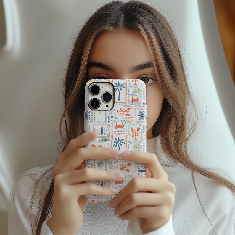 May include: A white phone case with a colorful, postage stamp-style design featuring crabs, fish, palm trees, and other nautical elements. The case is held up in front of a person's face.