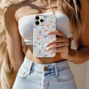 May include: A white iPhone case with a colorful, repeating pattern of stamps featuring palm trees, crabs, fish, and lobsters. The case has a protective design and is held in front of a person.