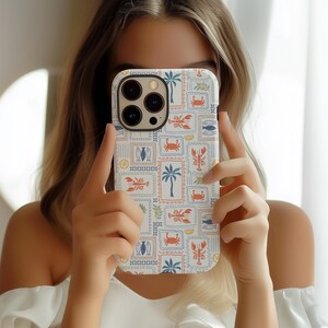 May include: A white iPhone case with a colorful, nautical-themed pattern. The design features postage stamp-like squares with illustrations of palm trees, crabs, lobsters, fish, and surfboards. The case has a protective bumper around the camera.