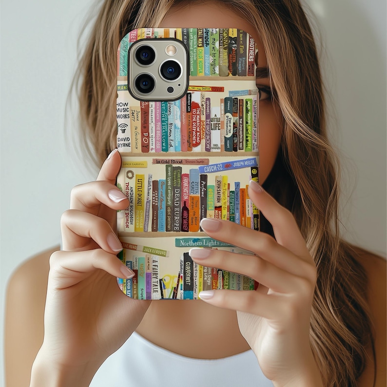 May include: A phone case featuring a bookshelf design with various book titles in multiple colors. The case has a beige background and is held in front of a person's face.