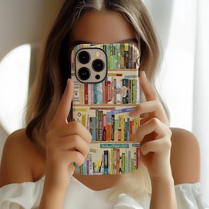 May include: A phone case featuring a colorful bookshelf design. The case displays rows of books with various titles and spine colors. The case has a black camera module and is held in front of a person.