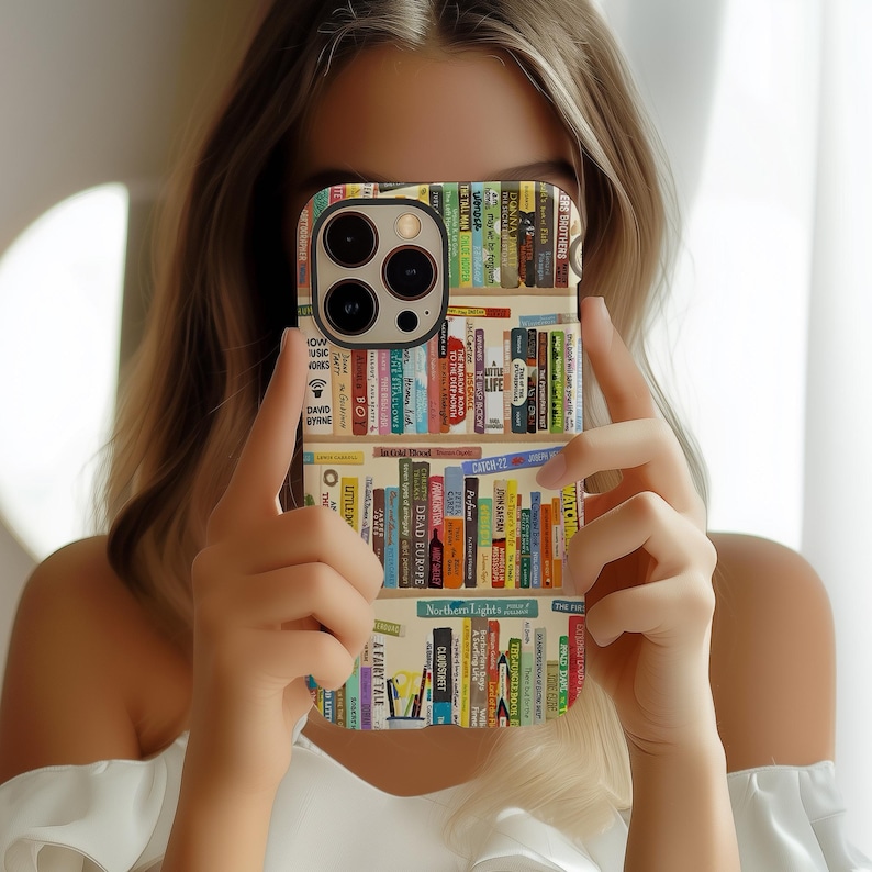 May include: A phone case featuring a colorful bookshelf design. The case displays rows of books with various titles and spine colors. The case has a black camera module and is held in front of a person.